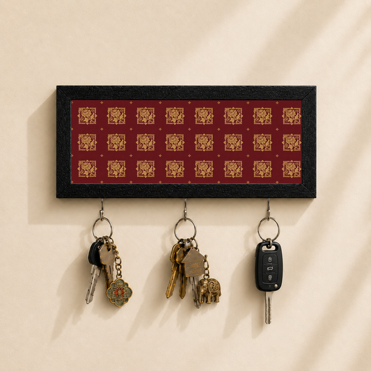 A rectangular black key holder with a red background and repeating gold decorative motifs, mounted on a beige wall. Three metal key rings hang below, holding keys and a car key fob.