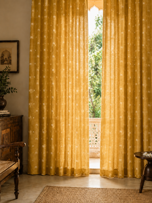 A pair of golden-yellow sheer curtains with white floral prints hanging on a window in a styled room with a wooden bench, side table, and a view of green trees outside.