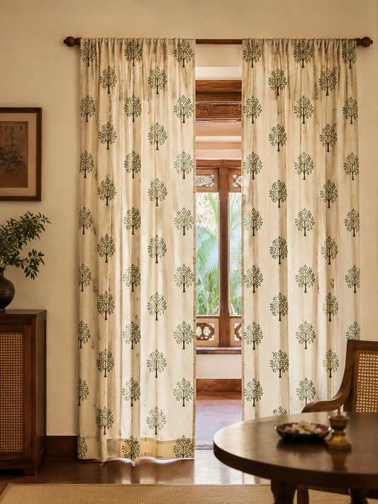 A pair of cream-colored curtains with a repeating green tree block print, hanging on a wooden rod in a warm, traditional room setting.