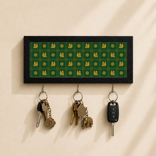 A rectangular wall-mounted key holder with a black border and a green patterned center panel featuring gold motifs. Three metal key rings hang below, holding keys and a car key, against a warm beige wall.