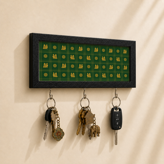 A rectangular black-framed wall key holder with a green and gold patterned insert, hanging on a beige wall. Three metal hooks below hold keys and a car key fob.