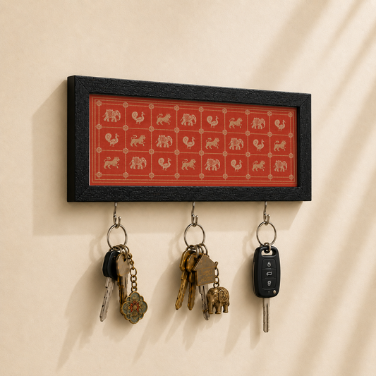 A rectangular black-framed wall key holder with a red-orange background and beige elephant motifs, mounted on a beige wall. Three metal hooks hang below, holding keys and a car key fob.