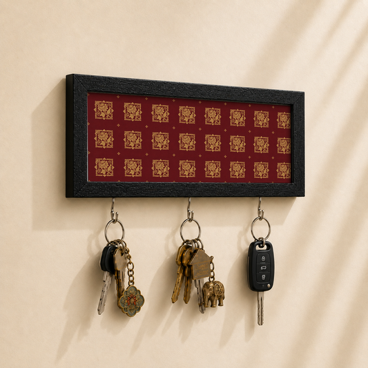 A rectangular black-framed wall key holder with a red background and repeating gold decorative motifs, mounted on a beige wall. Three metal hooks hang below, holding keys and a car key fob.