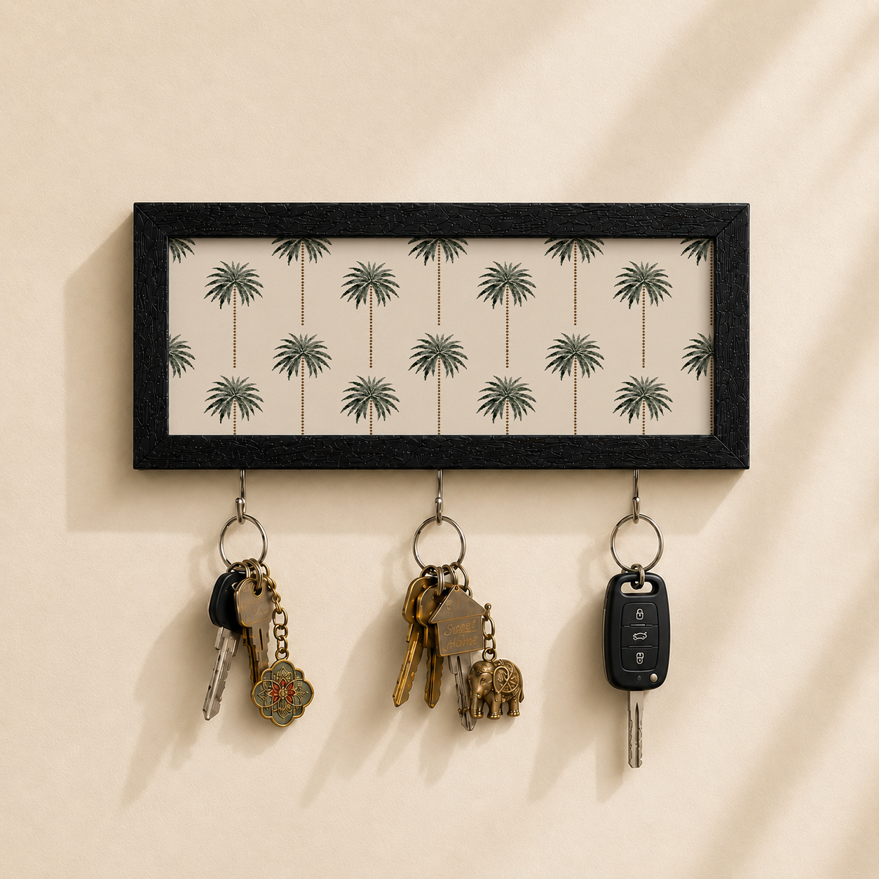 A rectangular black-framed wall key holder with a cream background and a repeating palm tree pattern, mounted on a beige wall. Three metal hooks hang below, holding keys and a car key fob.