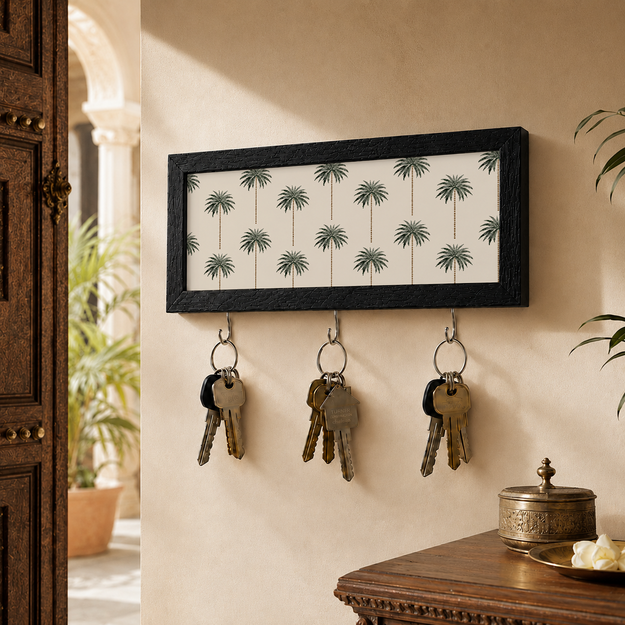A rectangular black-framed wall hook with a palm tree pattern on the front, hanging on a beige wall in a home interior. Three metal keys hang from the hooks, with a wooden door, plants, and a table visible in the background.