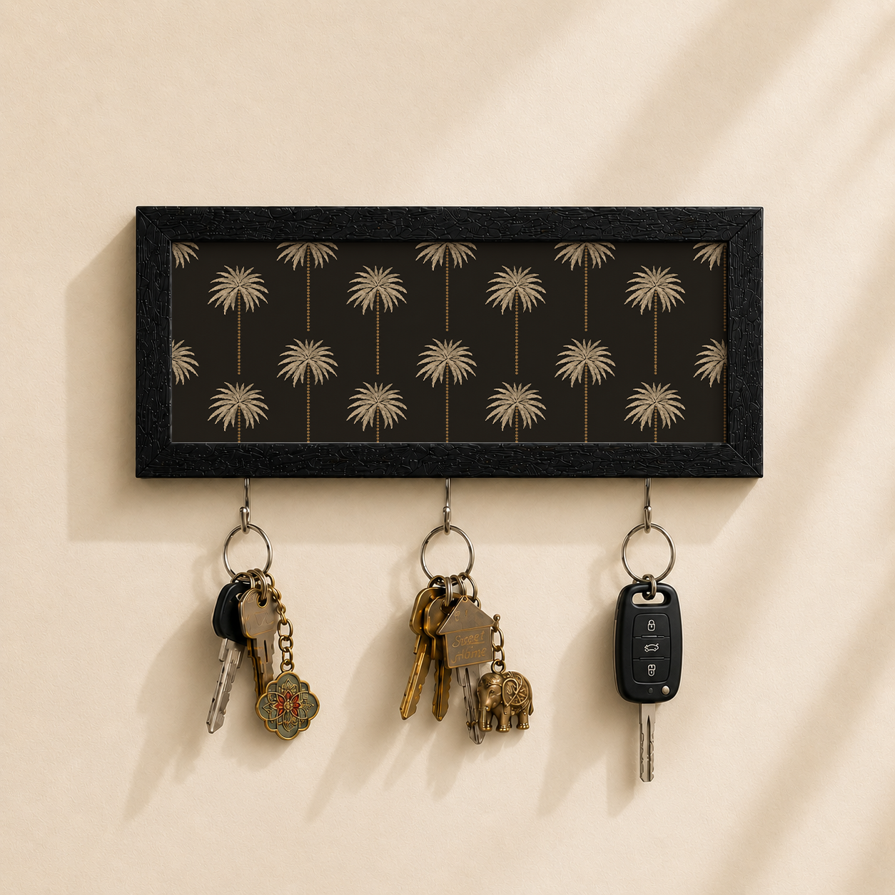A rectangular black wall-mounted key rack with a palm tree pattern, shown holding three sets of keys against a warm beige wall.