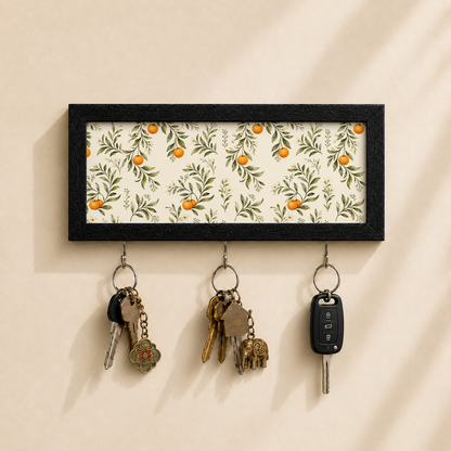 A rectangular black-framed wall key holder with a cream floral pattern featuring green leaves and orange fruits, hanging on a beige wall. Three metal key rings hang below, holding keys and a car key fob.