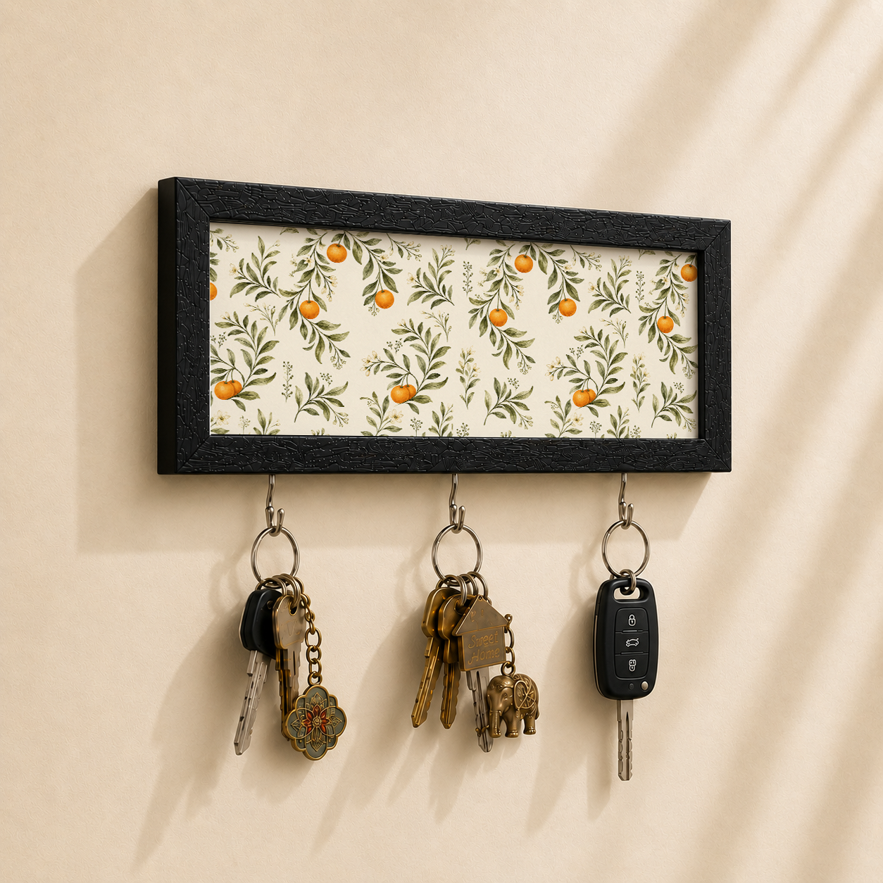 A rectangular black-framed wall key holder with a cream floral pattern featuring orange fruits, mounted on a beige wall. Three metal hooks hang below, holding keys and a car key fob.