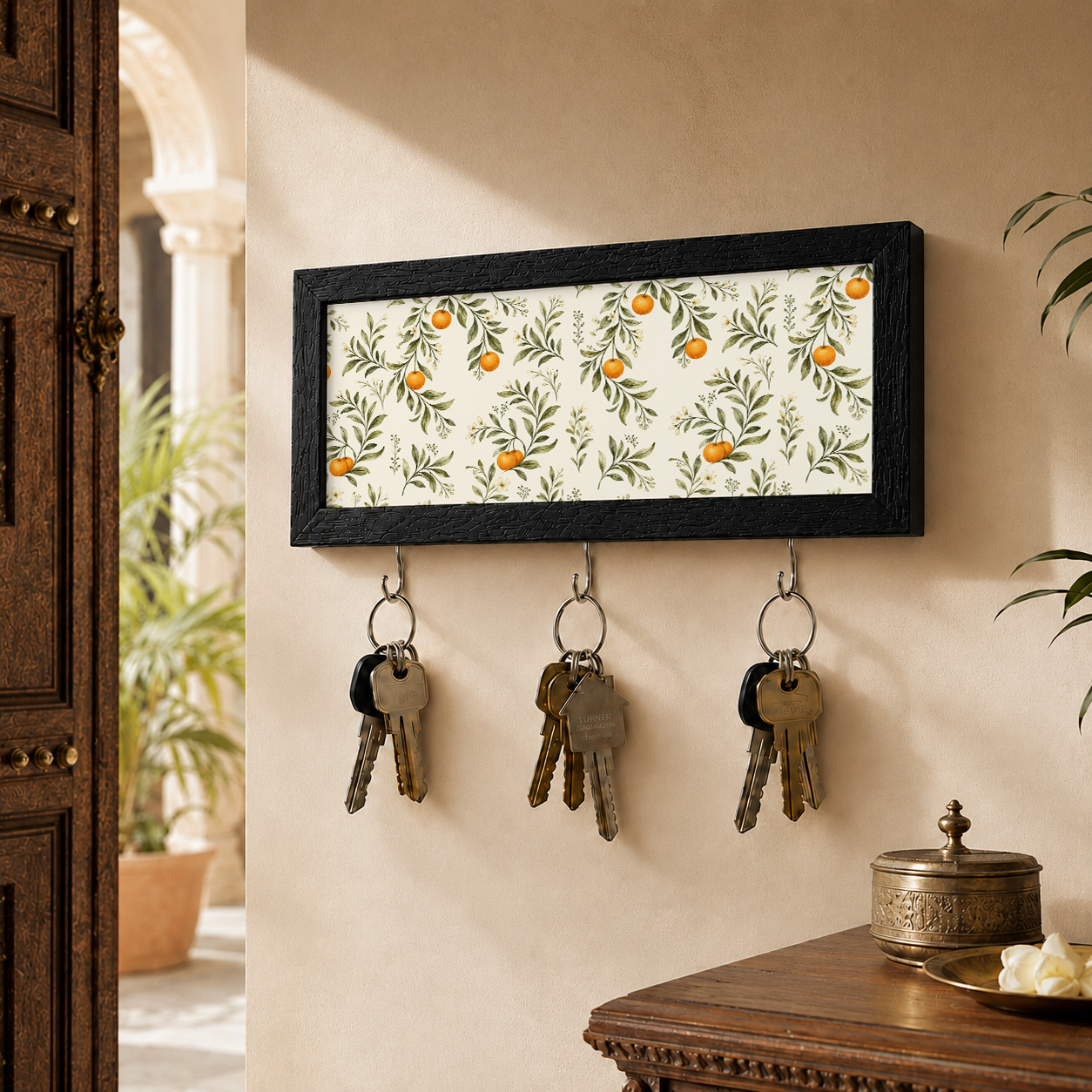 A rectangular black-framed key holder with a white background and an orange and green citrus pattern, hanging on a beige wall. Three metal hooks below hold keys, and part of a wooden door and table are visible in the room setting.