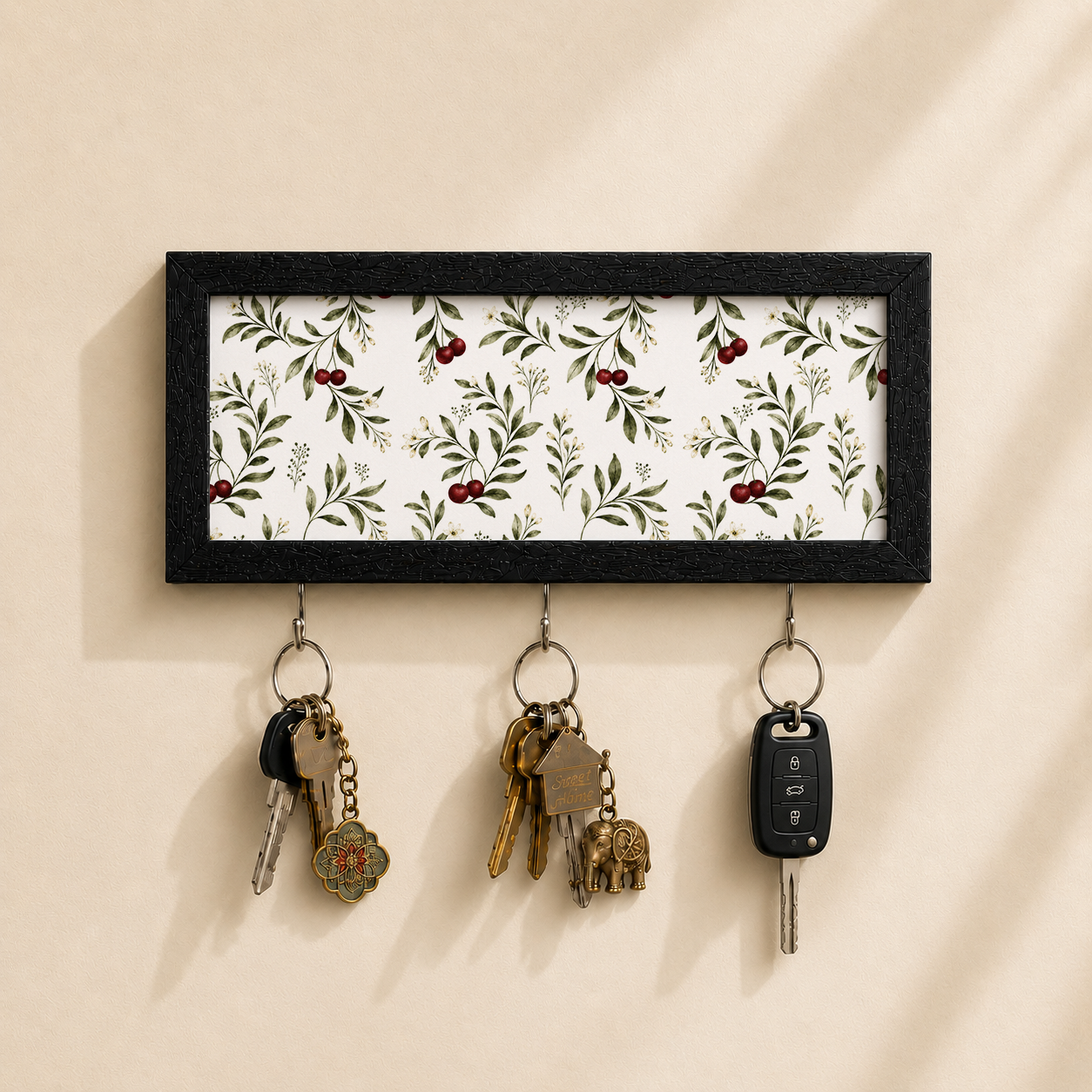 A rectangular black-framed key holder with a white background and a green leaf and red berry botanical pattern, mounted on a beige wall. Three metal key rings hang below, holding keys and a car key fob.