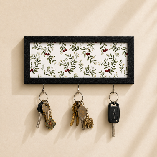 A rectangular black-framed key holder with a white background and a green leaf and red berry botanical pattern, mounted on a beige wall. Three metal key rings hang below, holding keys and a car key fob.