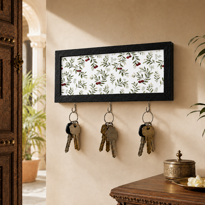 A rectangular black-framed key holder with a white background and a green and red berry botanical pattern, mounted on a beige wall. Three metal hooks hang below the frame, holding keys, with a wooden door and table visible in a styled interior setting.