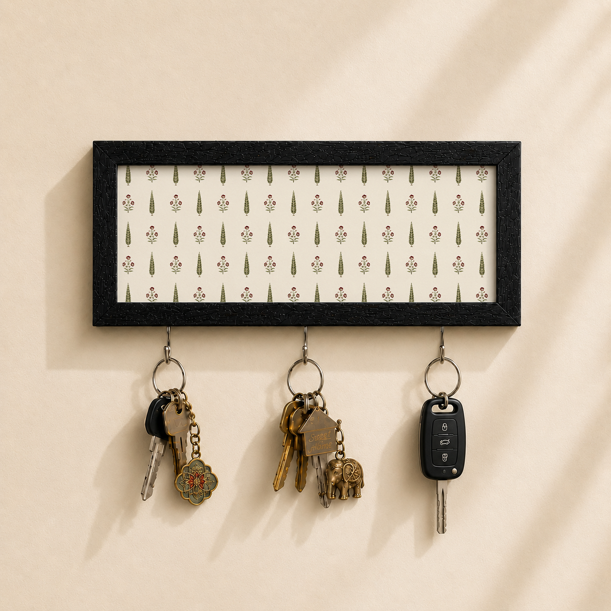 A rectangular black-framed wall key holder with a white patterned insert and three hanging metal keys, shown mounted on a beige wall.