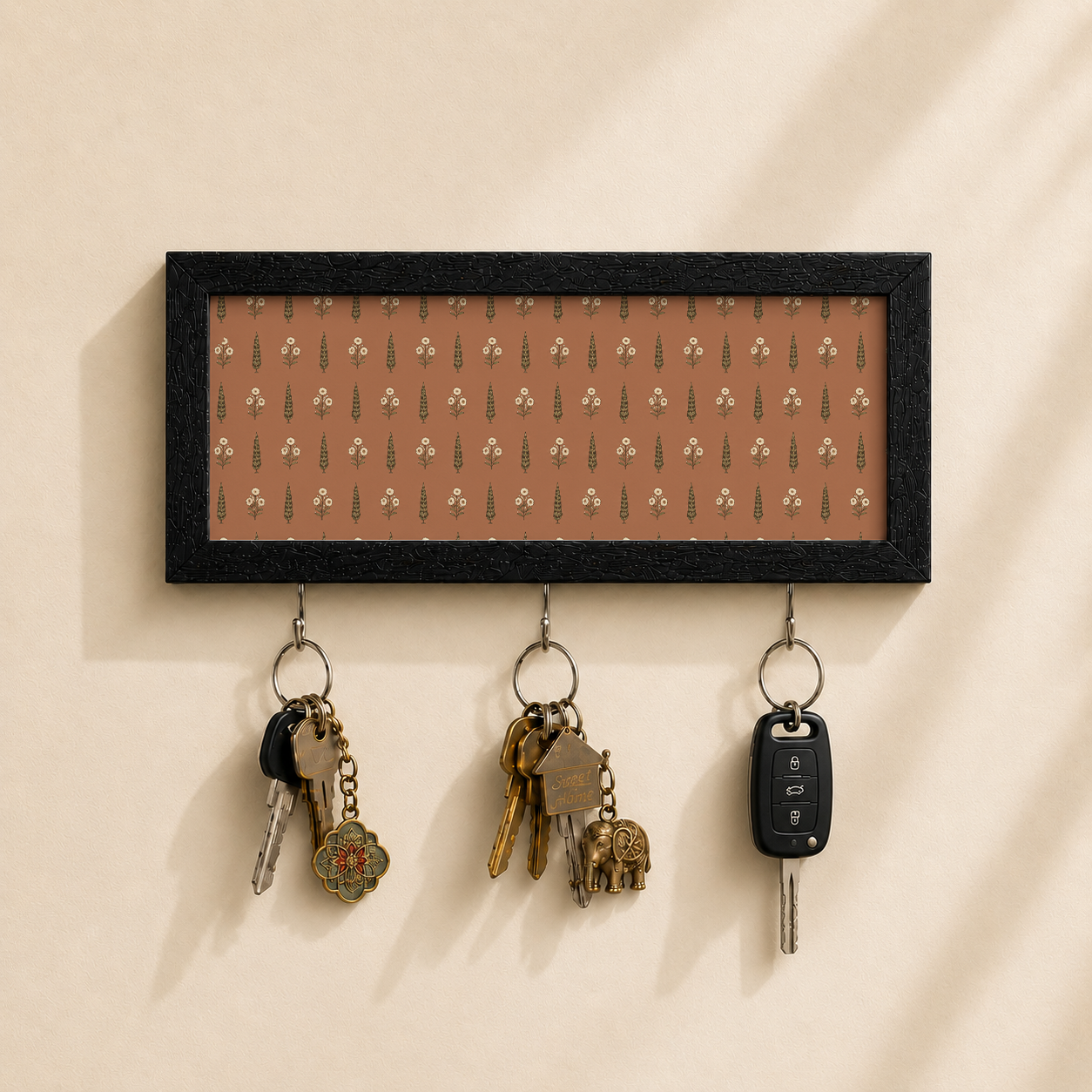 A rectangular black-framed wall key holder with a warm brown background and a repeating small floral pattern, mounted on a beige wall. Three metal hooks hang below, holding keys and a car key fob.