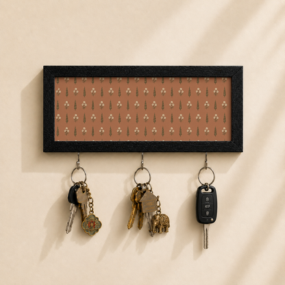 A rectangular black-framed wall key holder with a warm brown background and a repeating small floral pattern, mounted on a beige wall. Three metal hooks hang below, holding keys and a car key fob.