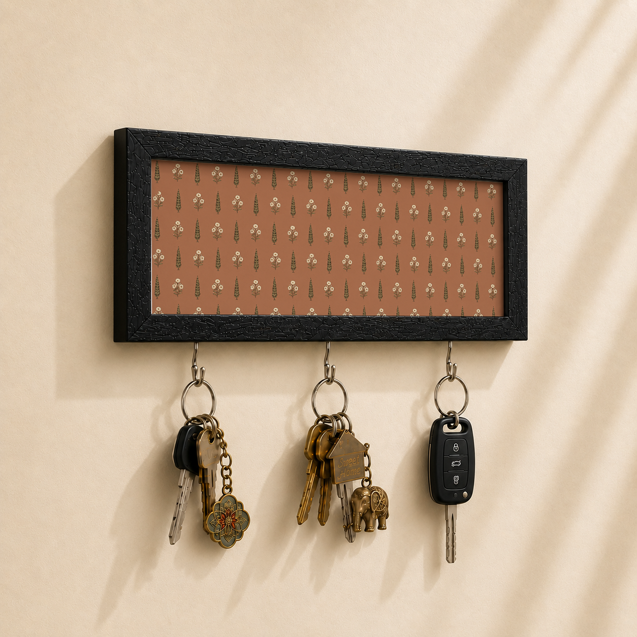 A rectangular black-framed wall hook with a warm brown patterned insert and three hanging metal keys, shown mounted on a beige wall with soft shadows.