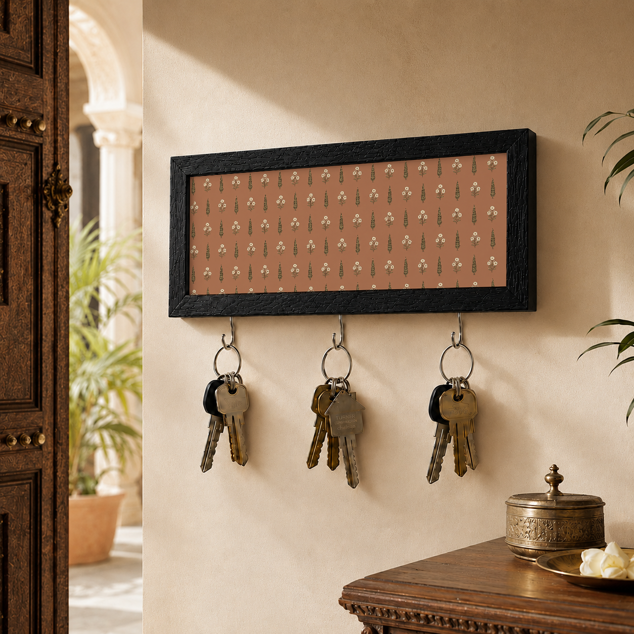 A rectangular black-framed wall key holder with a warm pink background and a repeating small floral pattern, mounted on a beige wall with indian home decor.