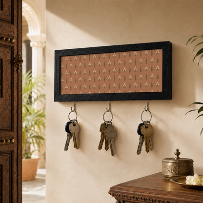 A rectangular black-framed wall key holder with a warm pink background and a repeating small floral pattern, mounted on a beige wall with indian home decor.