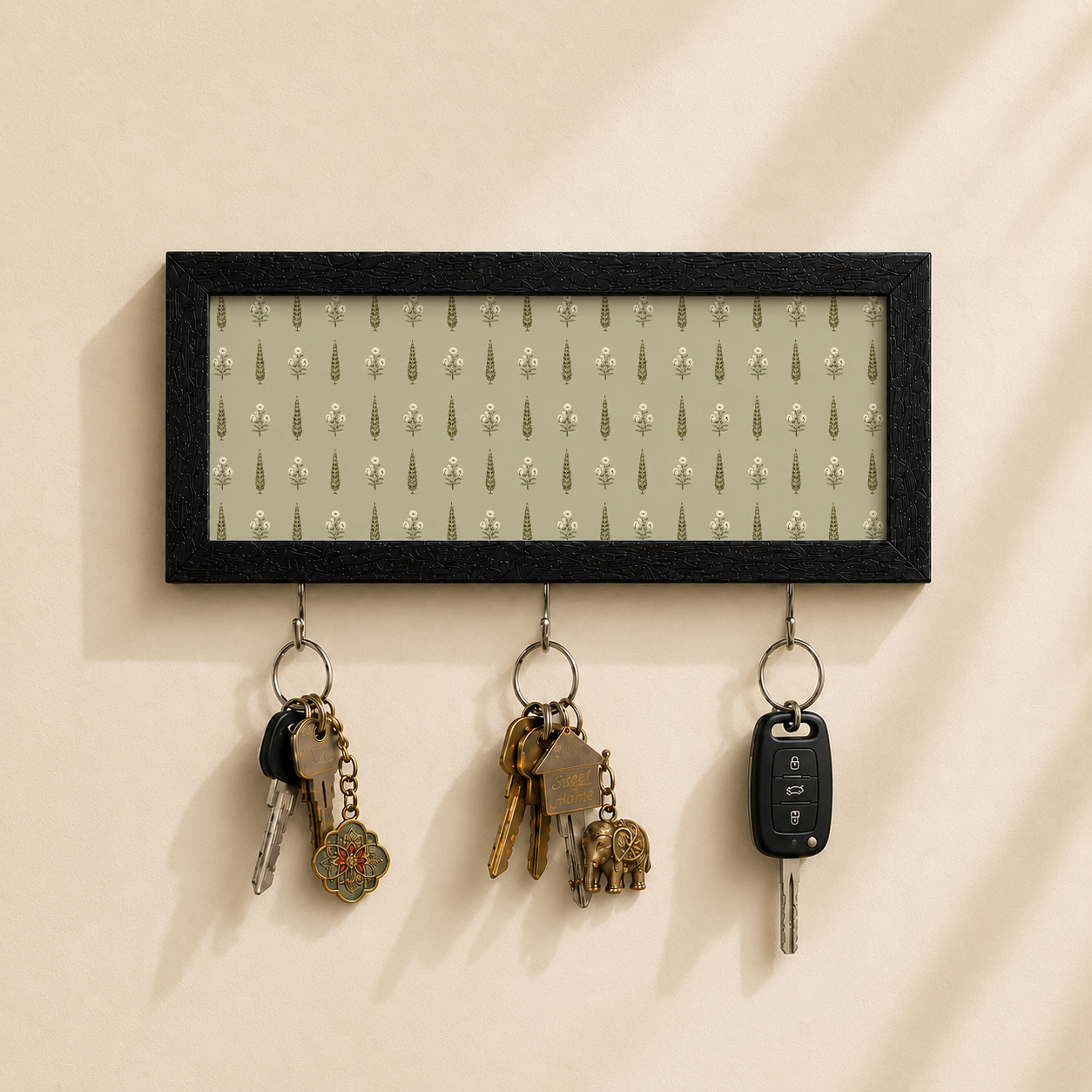 A rectangular black-framed wall key holder with a light sage green patterned insert, mounted on a beige wall. Three metal hooks hang below the frame, holding keys and a car key fob.