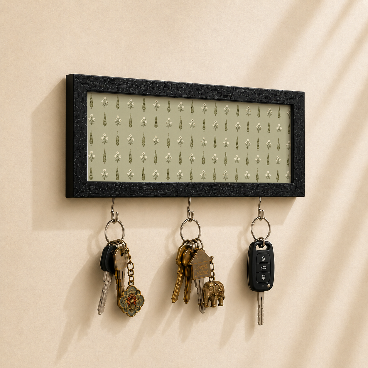 A rectangular black-framed wall key holder with a light sage green patterned insert, hanging on a beige wall. Three metal hooks below hold several keys and a car key fob.