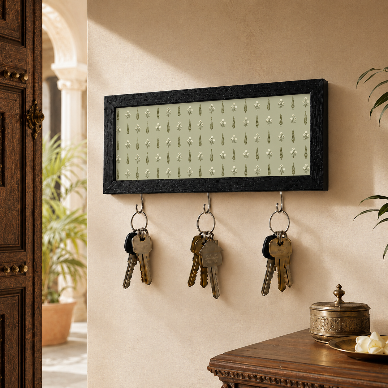 A rectangular black-framed key holder with a light green and white floral pattern, mounted on a beige wall. Three metal hooks hang below the frame, holding three sets of keys.