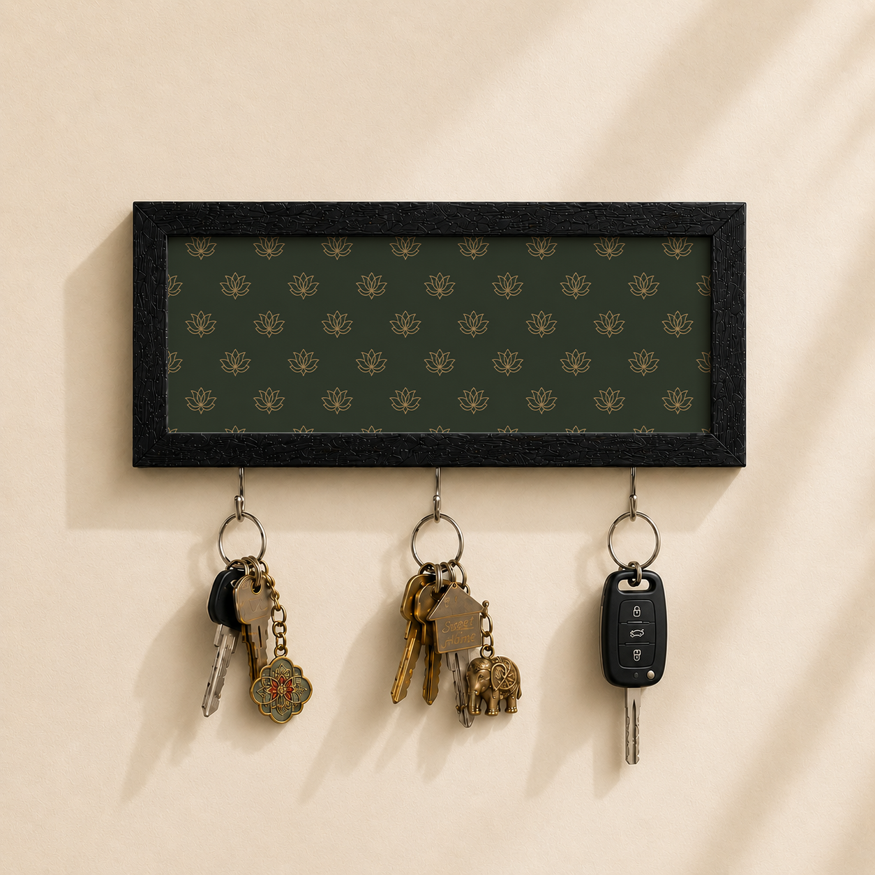 A rectangular black-framed wall key holder with a dark green patterned insert and three hanging keys, shown mounted on a beige wall with soft shadows.
