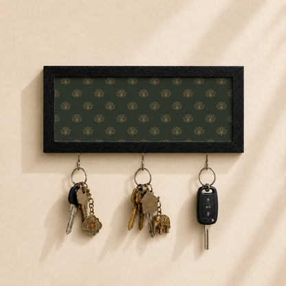 A rectangular black-framed wall key holder with a dark green patterned insert and three hanging keys, shown mounted on a beige wall with soft shadows.