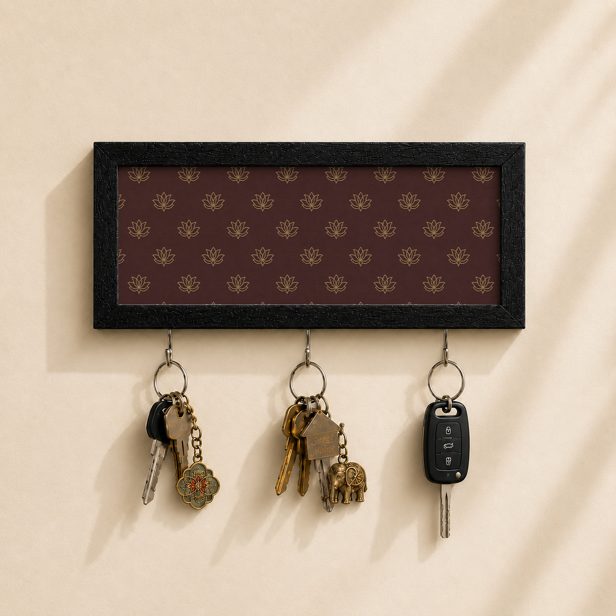 A rectangular black-framed key rack with a dark brown background and repeating gold floral pattern, mounted on a beige wall. Three metal hooks hang below, holding keys and a car key fob.