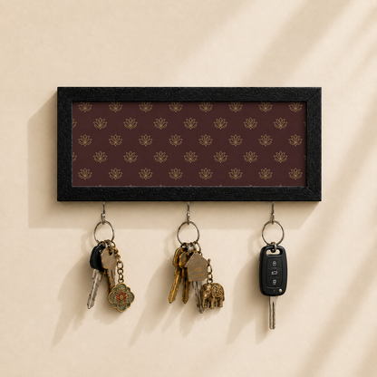 A rectangular black-framed key rack with a dark brown background and repeating gold floral pattern, mounted on a beige wall. Three metal hooks hang below, holding keys and a car key fob.