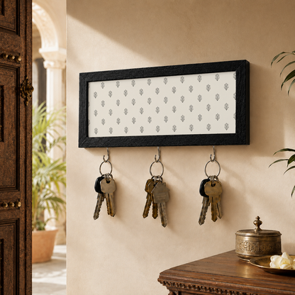 A rectangular black-framed wall hook rack with a white insert patterned with small gray motifs, mounted on a beige wall. Three metal hooks hang keys, and part of a wooden door and table are visible in the room setting.