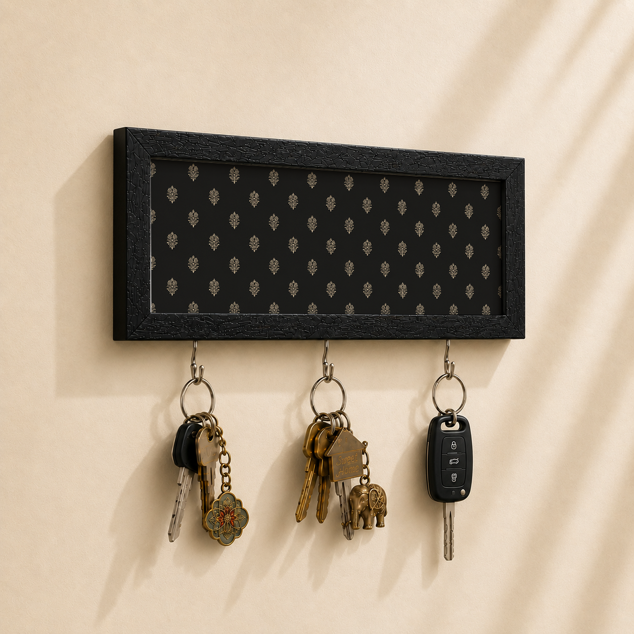 A rectangular black wall-mounted key holder with a subtle repeating floral pattern, shown mounted on a beige wall with three keys hanging from hooks below.