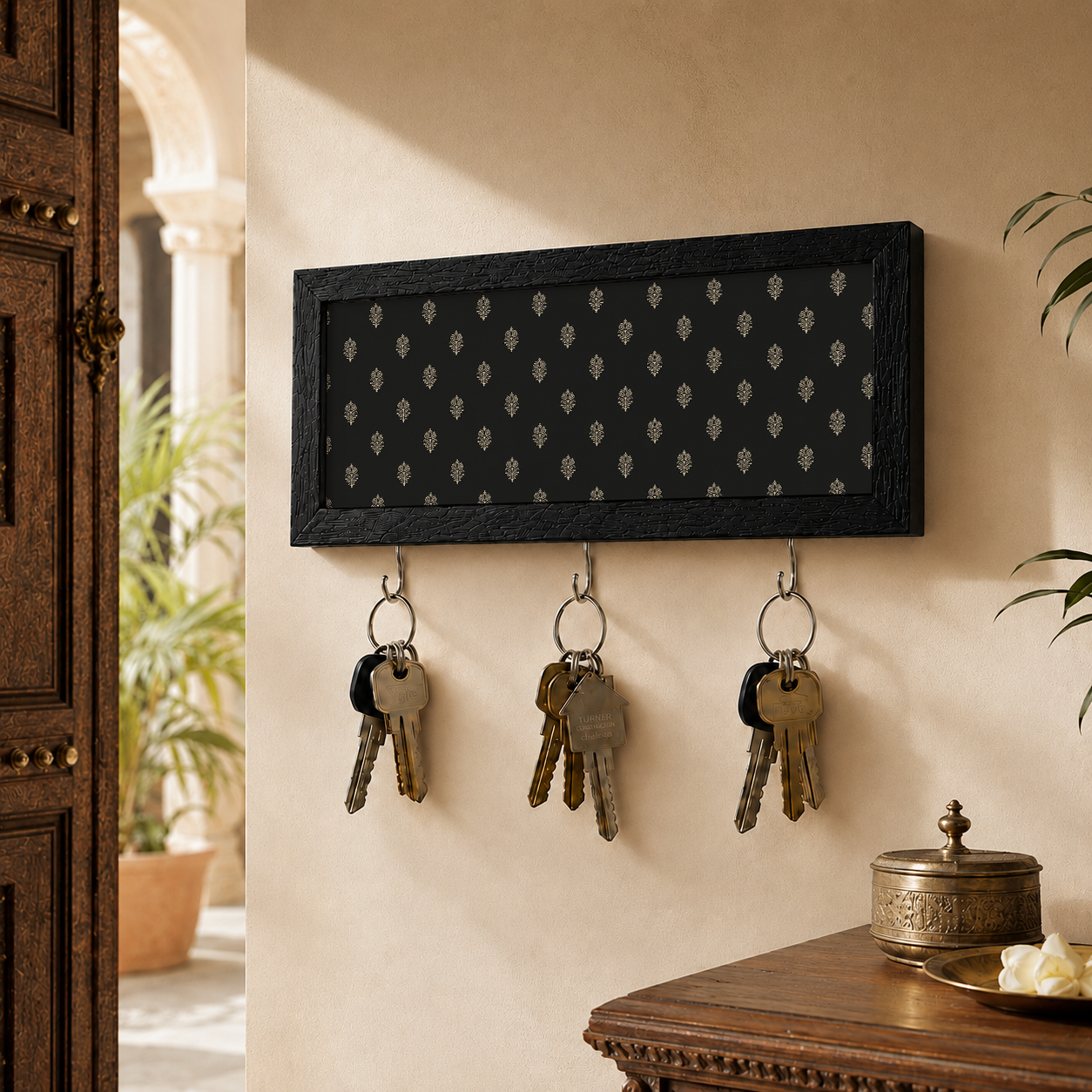 A rectangular black wall-mounted key rack with a small repeating light motif on the front and three metal hooks below, shown holding three sets of keys in a warm indoor setting with a wooden door, plants, and a table visible.