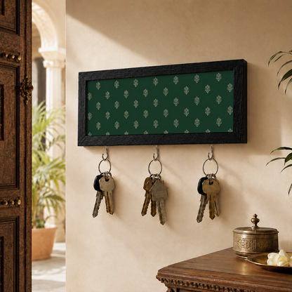 A rectangular wall-mounted key rack with a dark green damask-patterned insert and a black frame, hanging on a beige wall in a home entryway. Three metal keys are suspended from the hooks below.