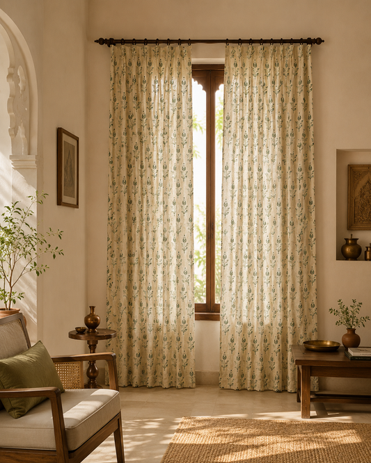 A pair of floor-length curtain panels with a delicate green floral vine pattern on a warm cream background, hanging on a dark rod in a beautifully styled room with warm beige walls, wooden furniture, and natural light.