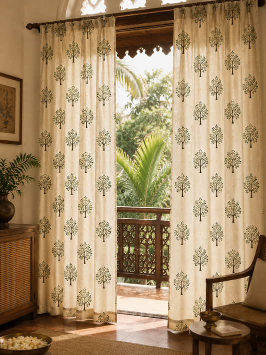 A pair of cream-colored curtains with a repeating green tree pattern, hanging on a dark wooden rod in a sunlit room with a balcony and tropical greenery visible outside.
