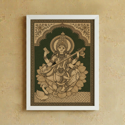 A framed Indian folk art print of the goddess Saraswathi seated on a lotus, displayed on a textured beige wall. The print features intricate tan and gold details on a dark green background, enclosed in a simple white frame.