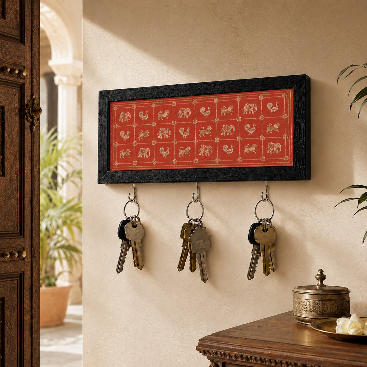 A rectangular wall-mounted key holder with a black frame and a red center panel featuring small animal silhouettes, hanging three keys on metal hooks in a warm indoor setting.