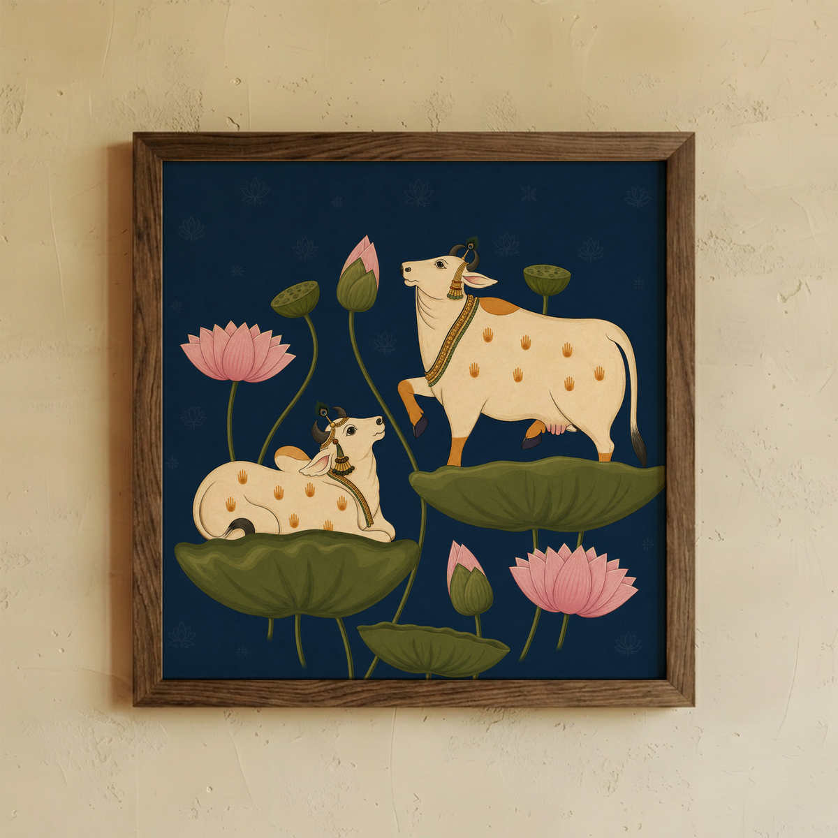 A square framed art print featuring a stylized illustration of two cream-colored spotted cows among green lotus pads and pink lotus flowers on a dark navy background, mounted on a textured beige wall.
