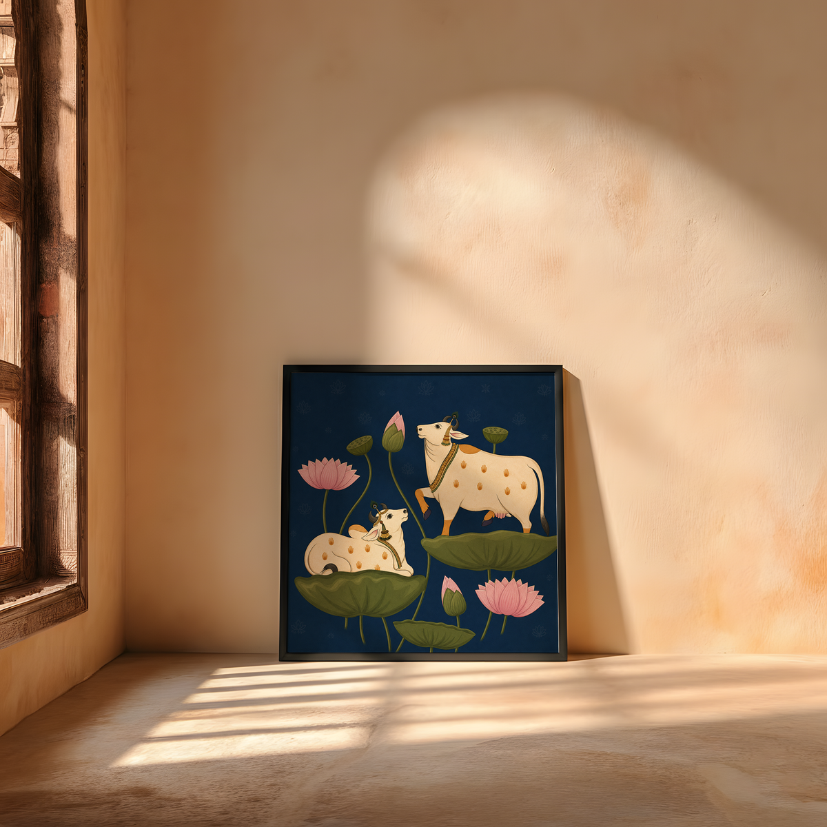 A framed art print featuring a stylized illustration of a cow and calf among lotus leaves and pink flowers, leaning against a warm beige plaster wall in a sunlit room.