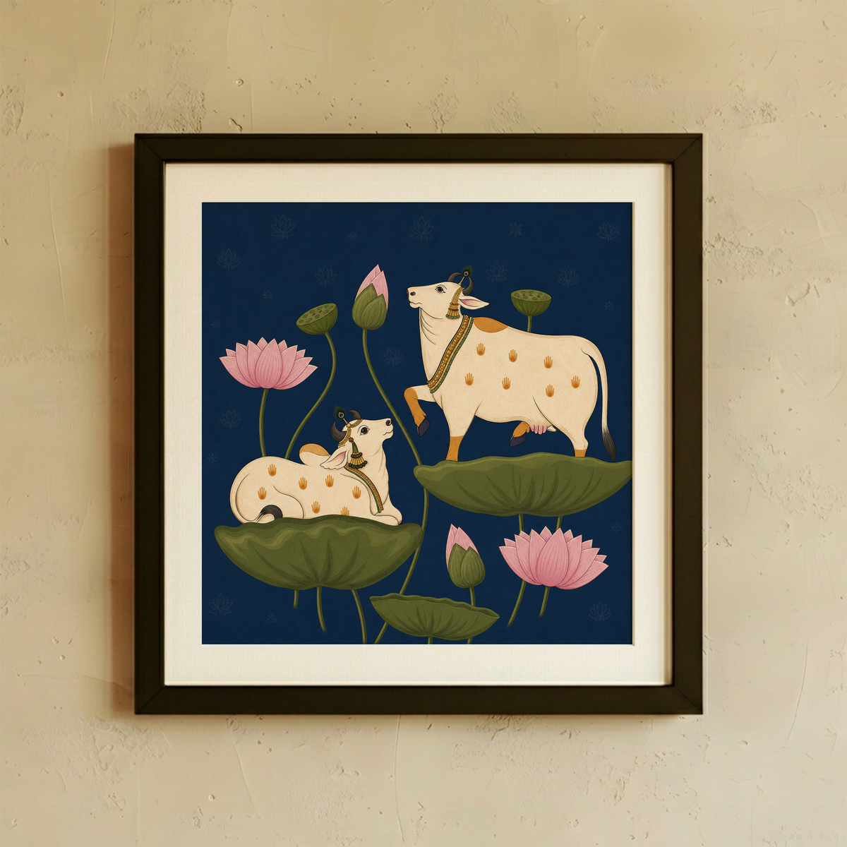 Framed wall art featuring an illustrated cream-colored cow and calf resting on green lotus leaves with pink lotus flowers against a dark navy background, displayed on a textured beige wall.