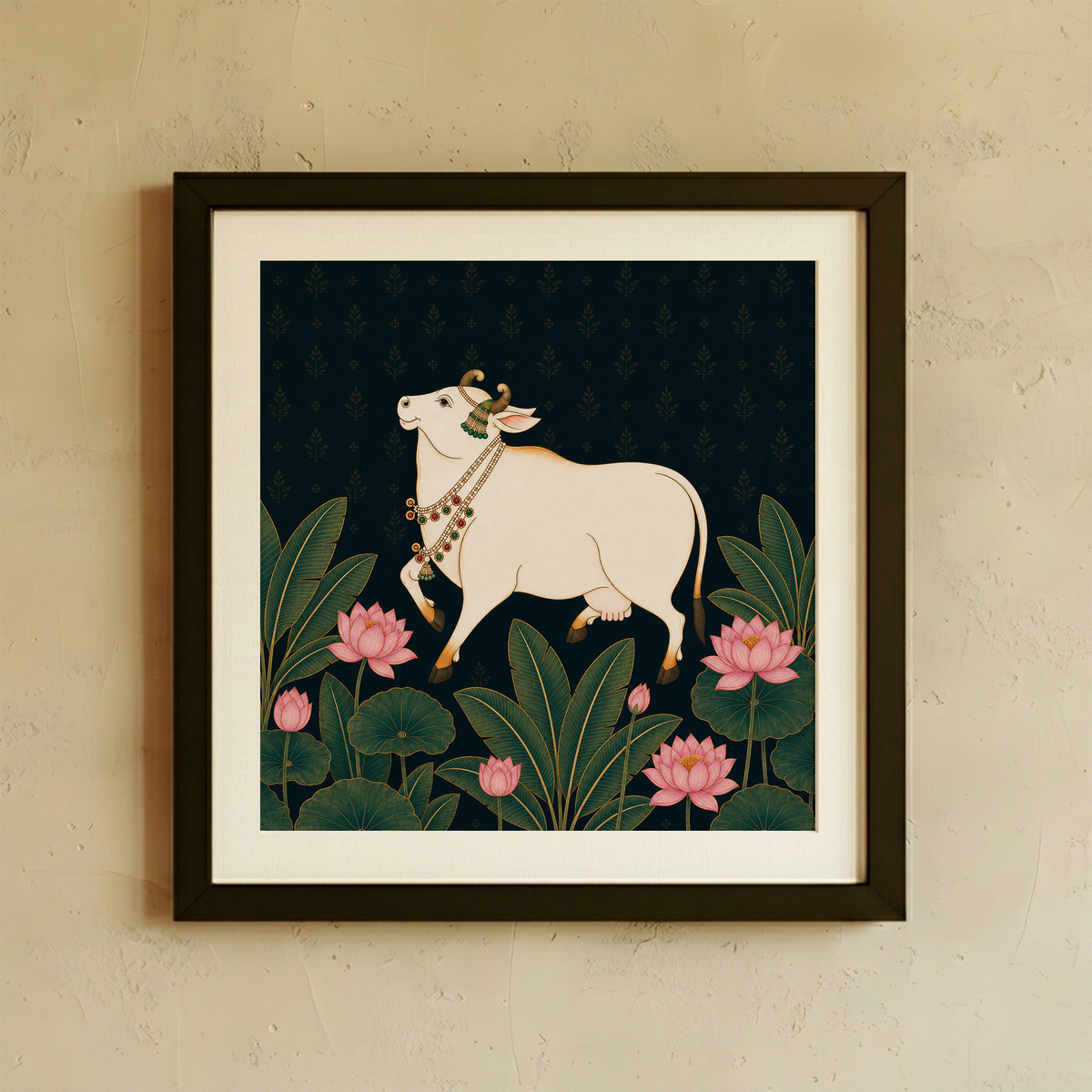 Framed wall art featuring a stylized white cow with decorative necklaces walking among green lotus leaves and pink lotus flowers on a dark navy background, shown hanging on a beige textured wall.