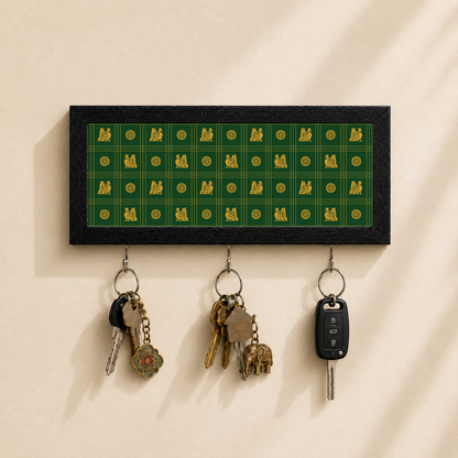 A rectangular wall-mounted key holder with a black border and a green patterned center panel featuring gold motifs. Three metal key rings hang below, holding keys and a car key, against a warm beige wall.