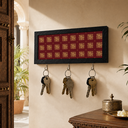 A rectangular wall-mounted key holder with a black frame and red background, decorated with small gold motifs. Three metal hooks hang below, holding keys, and the holder is shown mounted on a beige wall in a home entryway setting.
