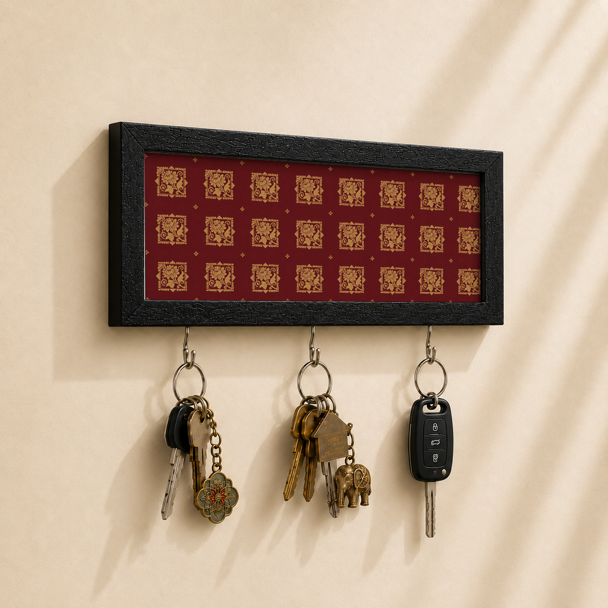 A rectangular black-framed wall key holder with a red background and repeating gold decorative motifs, mounted on a beige wall. Three metal hooks hang below, holding keys and a car key fob.