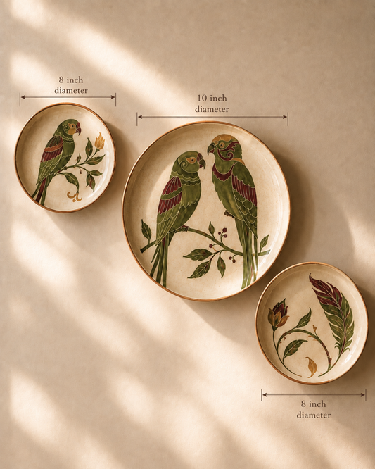 Three decorative ceramic plates with parrot and botanical illustrations, shown with dimension arrows and text indicating 8 inch and 10 inch diameters against a warm beige background with soft window shadows.