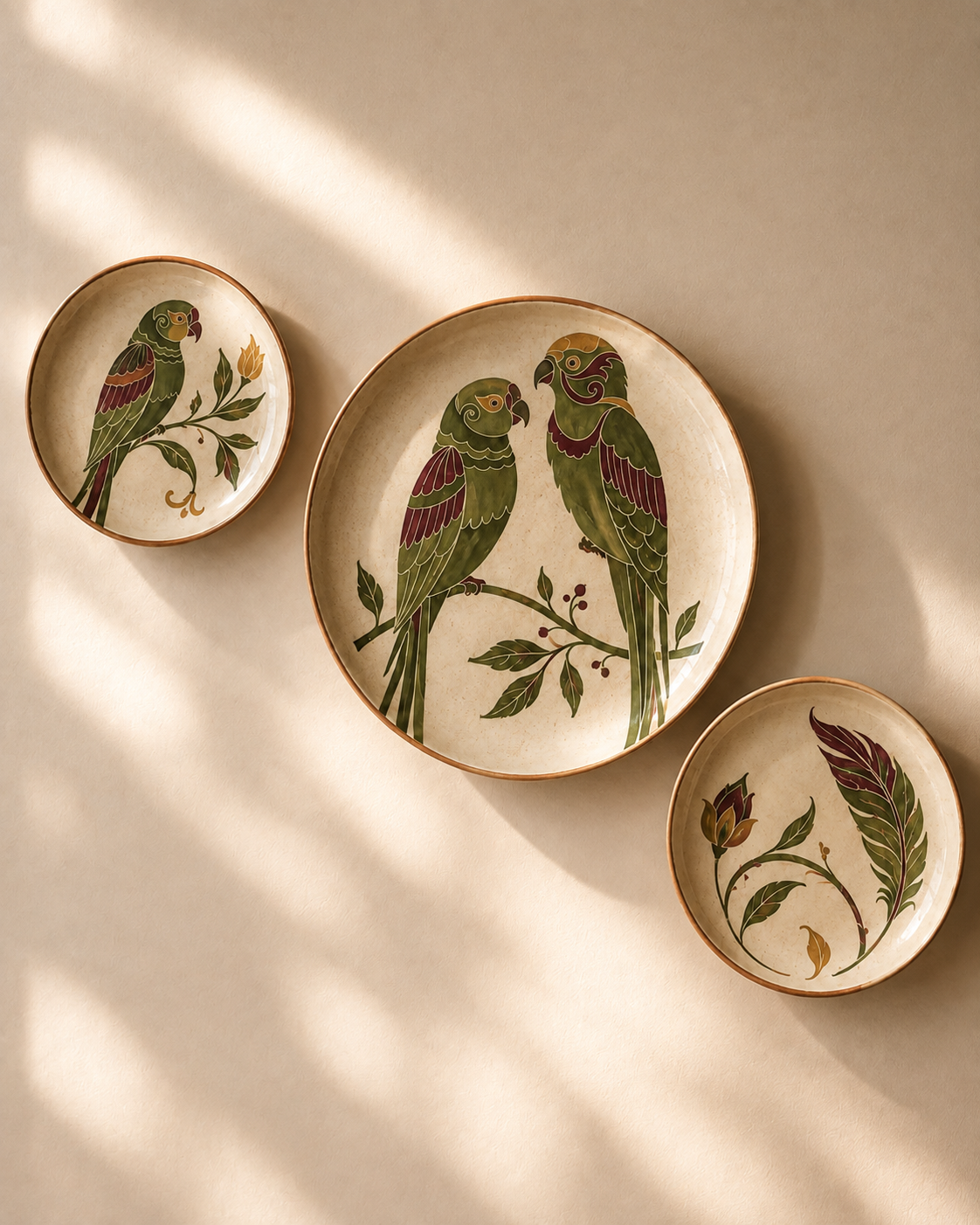 Three round ceramic wall plates with gold-toned rims are displayed on a beige wall. The central plate is larger and features two green parrots perched on a branch with small red berries.